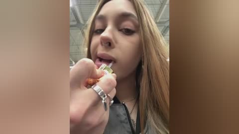 Media: Video of a young woman with long blonde hair, wearing a black top, eating a yellow snack with a silver ring. Background features a beige ceiling with metallic panels.