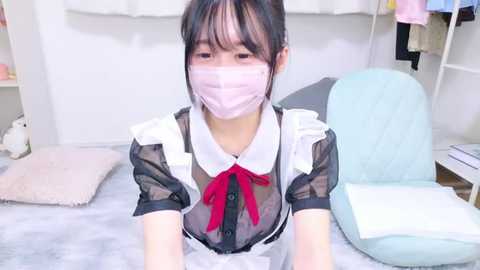 Media: Video of an Asian woman with a light complexion, wearing a black and white maid outfit, including a mask, and a red bow tie, sitting on a white cushioned chair in a clean, brightly lit room.