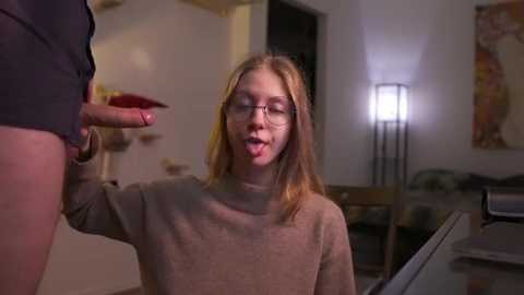 A video shows a blonde woman with glasses licking an erect penis, in a dimly-lit living room with modern art on the wall.