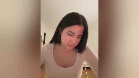 Media: Video of a young woman with medium skin tone, straight black hair, and a white long-sleeve shirt, sitting on a wooden floor, looking down with a soft smile, blurred background.