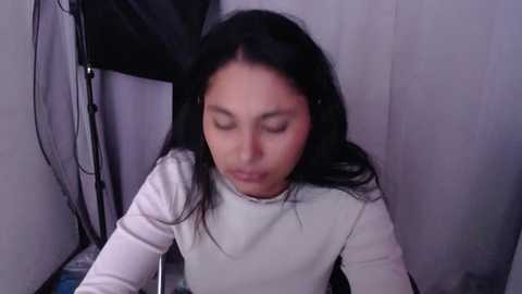 Media: Video of a young Asian woman with long black hair, wearing a white long-sleeve shirt, eyes closed, leaning forward, in a dimly lit room with a black mesh screen and white curtains.