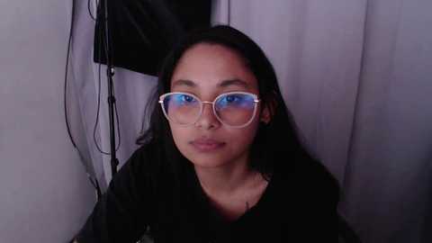 Media: Video of a young woman with long black hair, medium skin tone, wearing large, rectangular glasses and a black top, against a grey curtain backdrop.