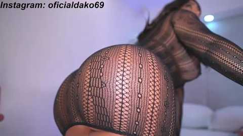 Video of a woman in black fishnet bodysuit, emphasizing her curvaceous buttocks and bare breasts. The background is a minimalist room with soft lighting.