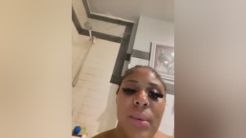 Media: A close-up video of a Black woman with dark skin, styled hair, and full makeup, including long eyelashes and pink lipstick, in a modern bathroom with white walls and a showerhead.