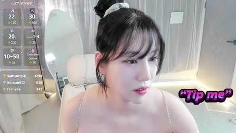 Media: Video of an East Asian woman with fair skin and black hair in a bun, wearing a white spaghetti-strap top, sitting in a modern, bright bathroom with white walls and a bathtub.