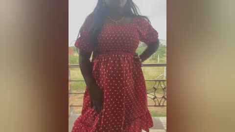 Media: Video of a dark-skinned woman with long black hair, wearing a red polka-dot dress with puffed sleeves, standing on a balcony with a blurred outdoor view.