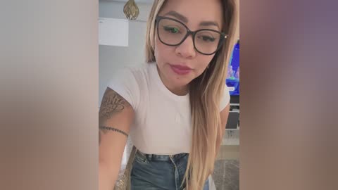 Video of a young, light-skinned woman with long blonde hair, wearing black-framed glasses and a white t-shirt, leaning forward, showing tattoos on her left arm.