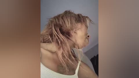 A video of a person with wet, tangled, pinkish-blonde hair, wearing a white, ribbed tank top, standing in a dimly lit room with neutral-colored walls.