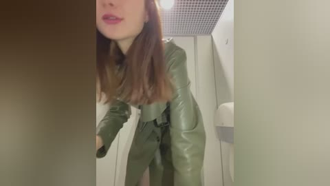 Media: A video of a woman in a green leather trench coat, her face partially obscured by shadows, standing in a bathroom with a white sink and ceiling tiles.