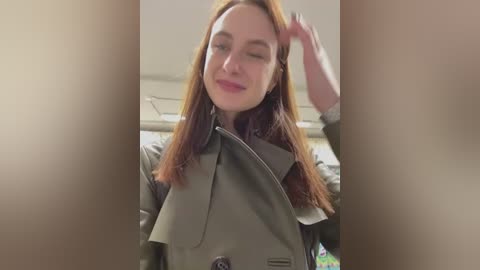 Media: Video of a smiling, light-skinned woman with long, straight, reddish-brown hair, wearing a green trench coat, indoors. She has her hand on her head, looking at the camera with a closed right eye.