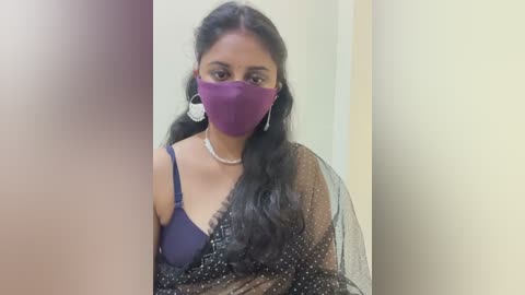 Media: Video of a South Asian woman with long black hair, wearing a purple mask, black sari with a blue bra visible, and pearl necklace, standing indoors against a plain wall.