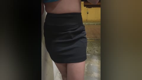 Media: Video of a light-skinned woman in a tight, black mini skirt, standing in a dimly lit doorway, revealing her midriff and legs, with a yellow wall and cobblestone path visible in the background.