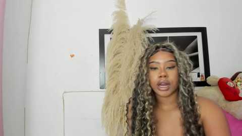 Media: Video of a Black woman with long, curly, gray and black hair, wearing a pink top, holding a large, beige ostrich feather. White wall, framed photo, red purse in background.