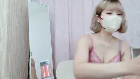 Media: Video of a young Asian woman with short blonde hair, wearing a pink lace bra and white face mask, sitting with arms crossed in a room with beige wallpaper and a mirror.