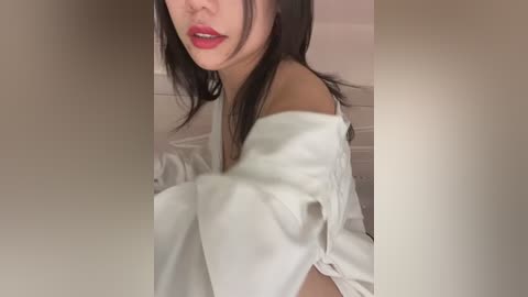 Media: Video of an Asian woman with long black hair, wearing a white off-shoulder top, lips painted red, looking slightly to the side, with blurred background.