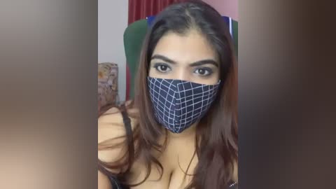 Media: Video of a young woman with long dark hair, wearing a blue and white checkered face mask, smiling, indoors with red curtains and a patterned cushion in the background.