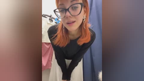 Media: A video of a fair-skinned woman with bright orange hair and black-framed glasses, wearing a black long-sleeve top and white skirt, leaning forward in a bathroom with a red checkered towel and blue shower curtain in the background.