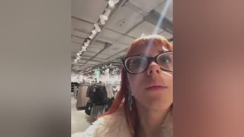 Media: Video of a pale-skinned woman with red hair, wearing black-rimmed glasses and a white fur coat, standing in a brightly lit, modern clothing store with hanging racks and fluorescent lighting.