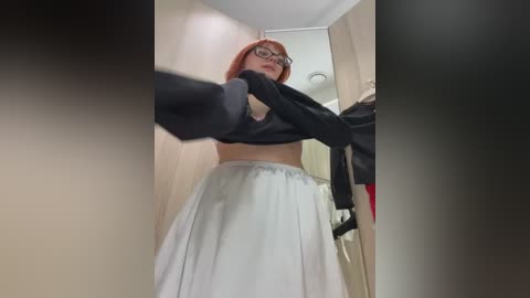 A video of a young woman with red hair, glasses, and a black top, lifting her skirt to reveal a white skirt, standing in a modern, well-lit hallway.