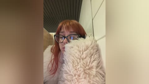 Video of a woman with long red hair, glasses, and a fluffy white coat, partially obscured by a fluffy white object, in a dimly lit corridor with a textured ceiling.