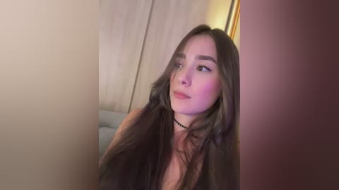 Video of a young Asian woman with long, dark hair, light skin, and a black choker necklace. She has a contemplative expression, with a wooden wardrobe and warm lighting in the background.