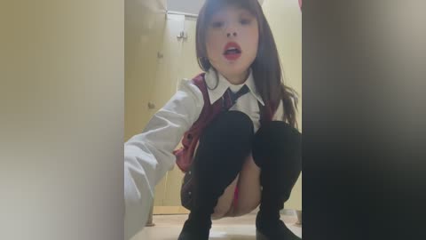 Media: Video of a young Asian woman in a schoolgirl uniform squatting, revealing red panties, in a hallway with beige walls and a door.