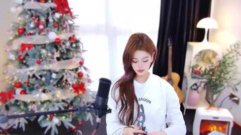 Media: Video of a young Asian woman with long brown hair, wearing a white T-shirt with a graphic, recording a video in a cozy room with a decorated Christmas tree and candles.