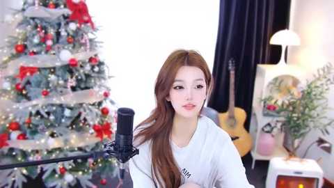 Media: Video of a young Asian woman with long brown hair, wearing a white T-shirt, singing into a microphone in a cozy, decorated room with a Christmas tree and guitar.