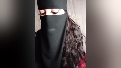 Media: Video of a person with long, dark hair, partially obscured by a black niqab, revealing only the eyes. The background is blurred, with a hint of a white wall.