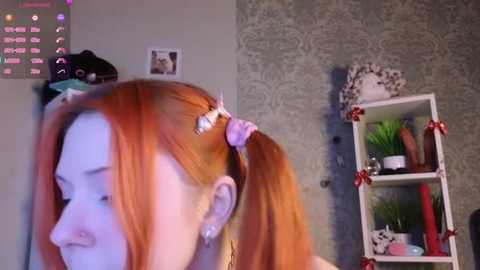 Video of a young woman with fair skin and vibrant orange hair styled in pigtails, adorned with pink scrunchies. She has a small nose piercing. Background features a light green wallpaper, a small white shelf with green plants and a stuffed animal, and a digital clock.