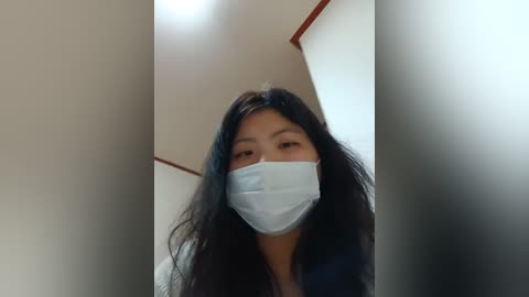 Media: A video of an East Asian woman with long, straight black hair, wearing a light blue surgical mask and a dark top, standing in a beige hallway with a white ceiling and brown trim.