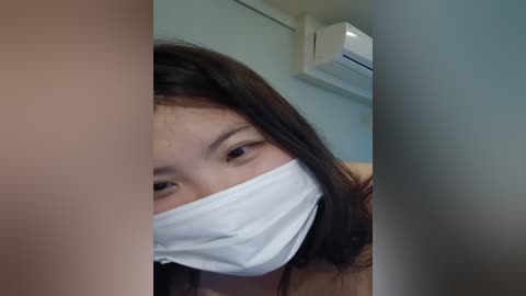 Media: Video of an Asian woman with straight dark hair, wearing a white surgical mask and a beige jacket, indoors with an air conditioning unit visible in the background.