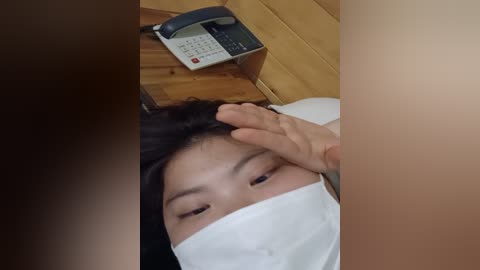 Media: A video shows a young woman with long black hair, wearing a white surgical mask, lying on a wooden table, with a hand touching her forehead. An old-fashioned phone sits on the table behind her.