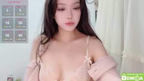 Media: Video of an East Asian woman with long, straight black hair, wearing a pale pink lace bra, off-shoulder jacket, and a neutral expression. Background includes a calendar and white curtain.