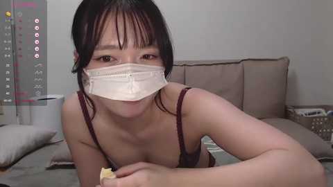 Media: A video of an Asian woman with short black hair and a white face mask, wearing a burgundy bra, lying on a bed with beige pillows and a white duvet.