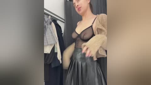 Media: A video of a light-skinned woman with short dark hair, wearing a sheer black top, beige cardigan, and high-waisted black leather skirt, standing in a changing room.