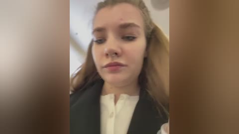 Media: Video of a young Caucasian woman with fair skin and long, blonde hair, wearing a black vest over a white blouse, looking tired with slightly puffy eyes. The background is out of focus, likely an indoor setting.