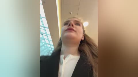 Media: Video of a Caucasian woman with long brown hair, wearing a black blazer over a white shirt, standing in a modern office with glass walls and fluorescent lighting.