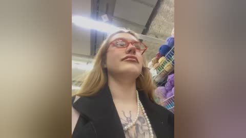 Video of a young woman with fair skin, long blonde hair, and red-framed glasses, wearing a black vest over a white graphic tee, standing in a yarn store with colorful skeins in the background.