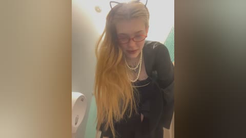 Media: Video of a young woman with long, straight blonde hair, wearing red glasses, a black top, and pearl necklace, leaning against a bathroom wall.