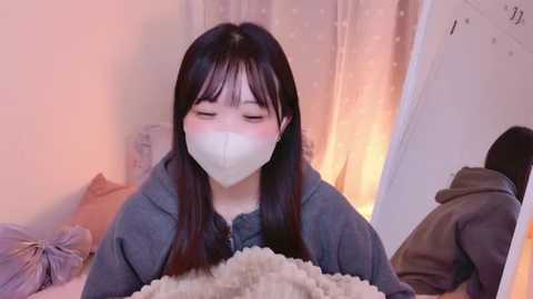 Media: Video of an Asian woman with long black hair, wearing a white face mask, gray hoodie, and holding a fluffy white cat. Soft, warm lighting. Bedroom background with beige curtains and a partially visible person in a dark hoodie.