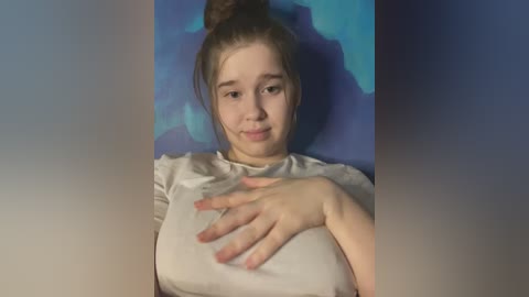 Media: A video of a young Caucasian woman with light skin, brown hair in a bun, wearing a light gray top, and resting her hand on her chest against a blue and purple backdrop.