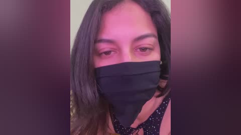 Media: Video of a woman with long dark hair, wearing a black face mask, a black top, and a beige background. Her brown eyes are partially visible.