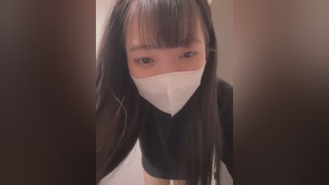 Media: A video of an East Asian woman with long black hair, wearing a white face mask, black top, and standing indoors.