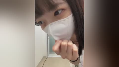 Media: Video of an Asian woman with long, dark hair, wearing a white surgical mask, peering through a doorway with a clenched fist. Background shows a plain, beige corridor with a closed door.