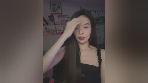 Media: Video of an East Asian woman with long, dark hair, wearing a black dress, wiping her forehead in a dimly lit room with a pink wall and shelves with toys.