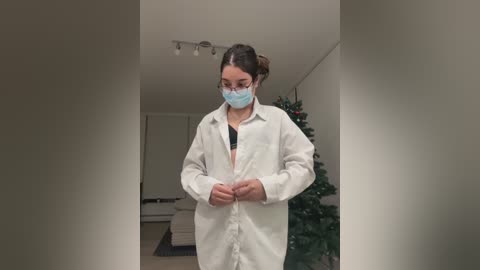 Media: Video of a young woman with light skin, brown hair in a bun, wearing a white lab coat, blue face mask, and black bra, standing indoors near a Christmas tree.