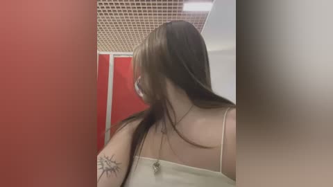 Media: A video of a woman with long brown hair and a tattoo on her shoulder, wearing a beige tank top, standing in a room with red and white walls and a grid-patterned ceiling.