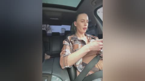 Video of a bald, white woman in a plaid shirt, driving a car with a safety belt on, viewed from the side.