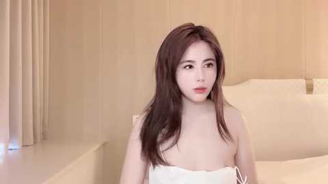 Media: Video of a young Asian woman with long, straight dark hair, wearing a white strapless dress, sitting on a bed in a minimalist, beige-toned room.